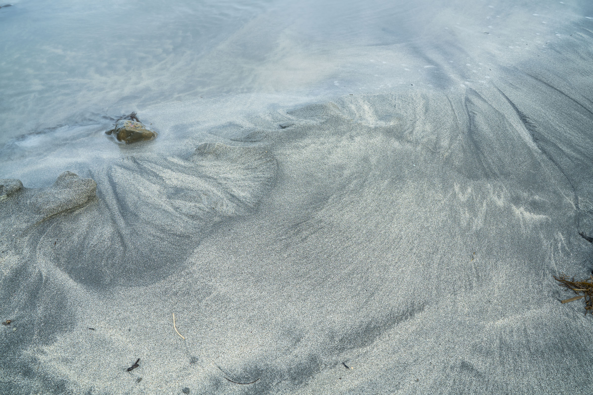 Water washing over rippled grey sand beside a small rock