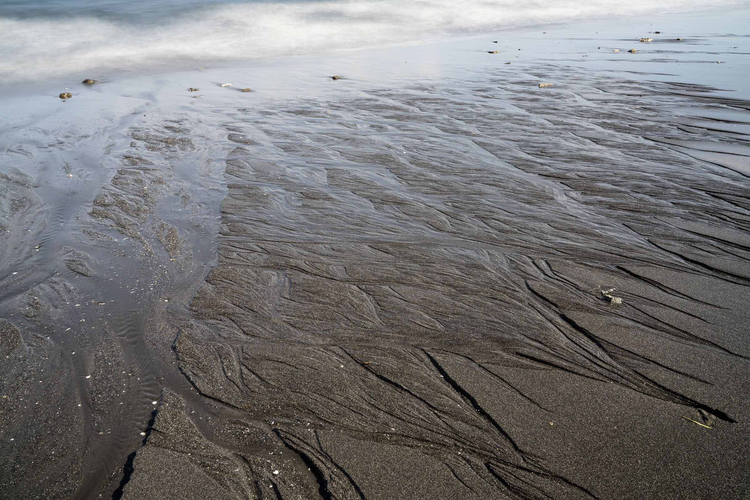 Branching rivulets across wet dark sand beside foamy water