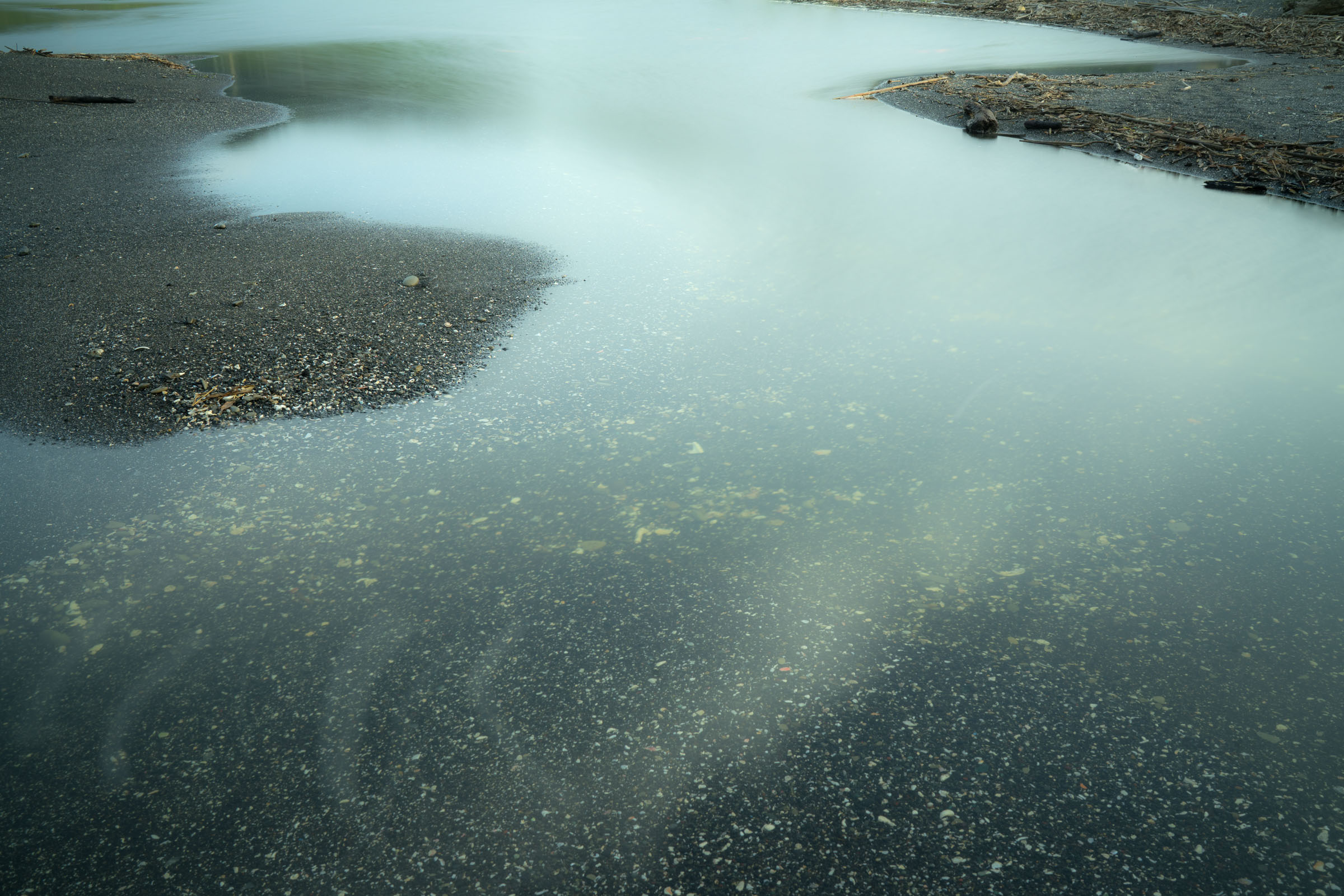 Pale water winding between dark pebbled banks and driftwood