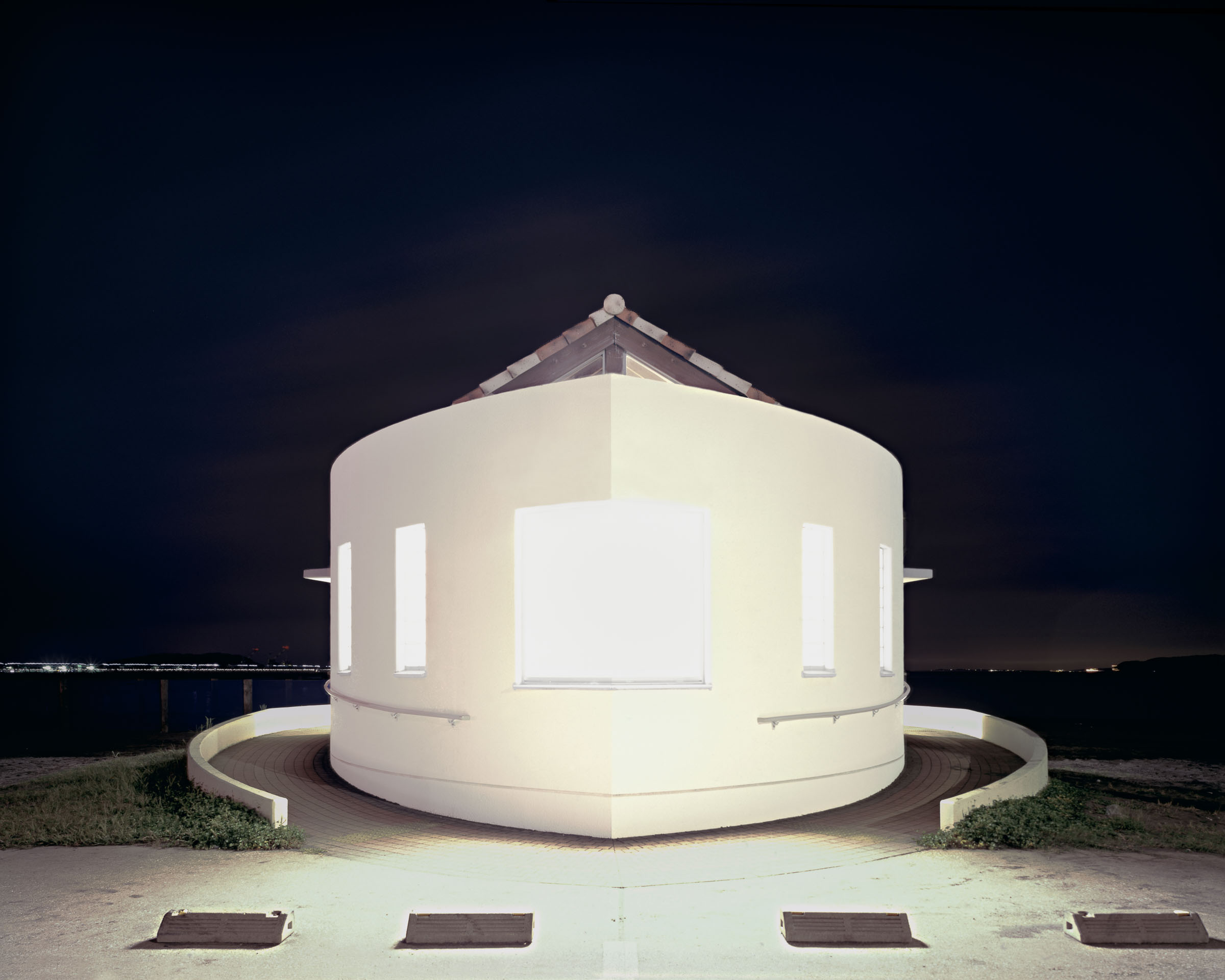 Round white public toilet with glowing windows beside the water