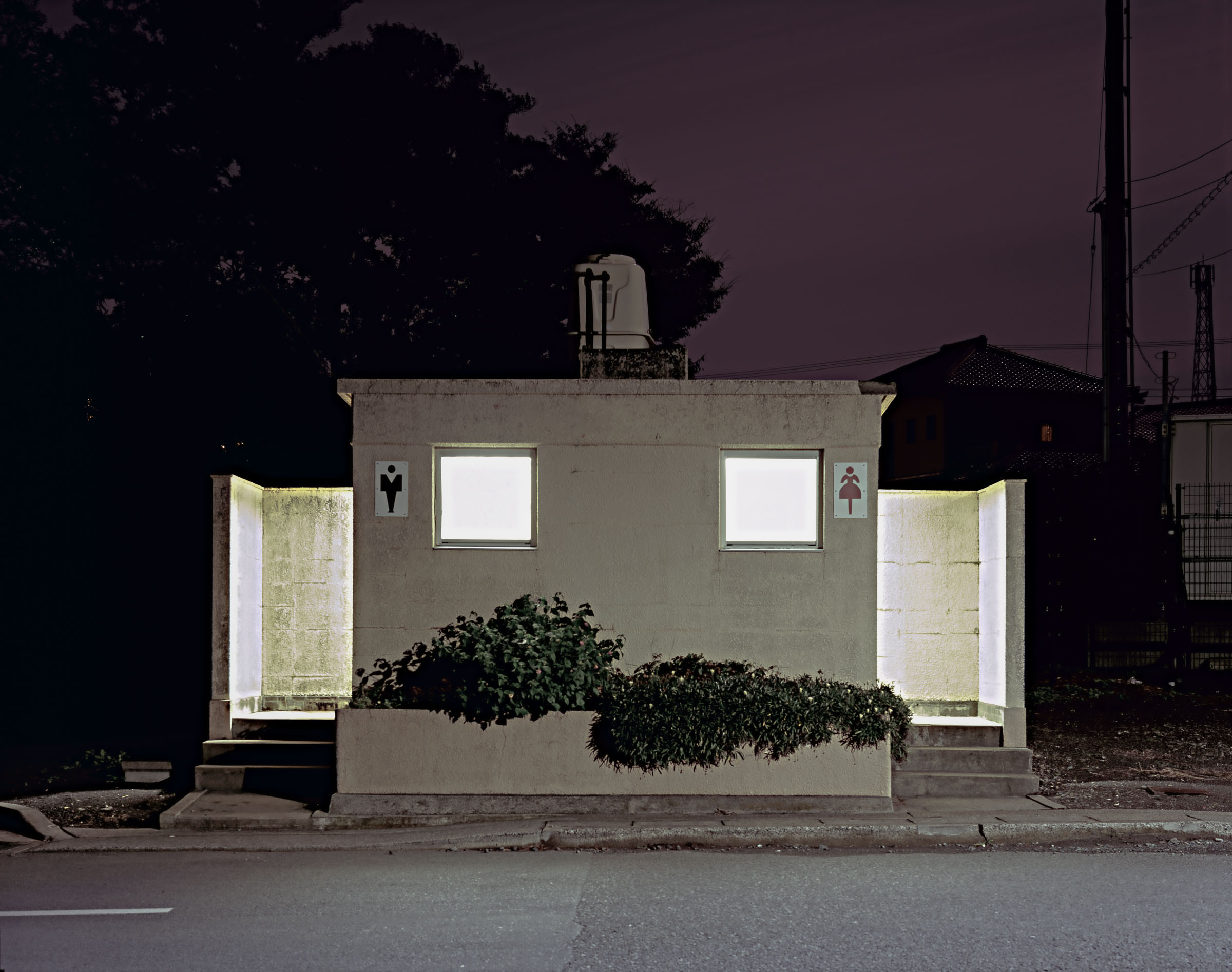 Rectangular white public toilet with glowing windows beside trees