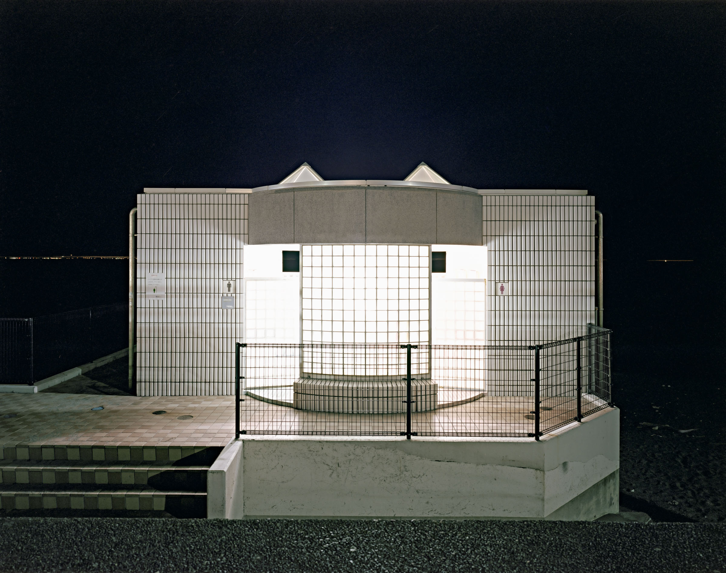 Tiled public toilet with an illuminated glass-block wall and metal fence