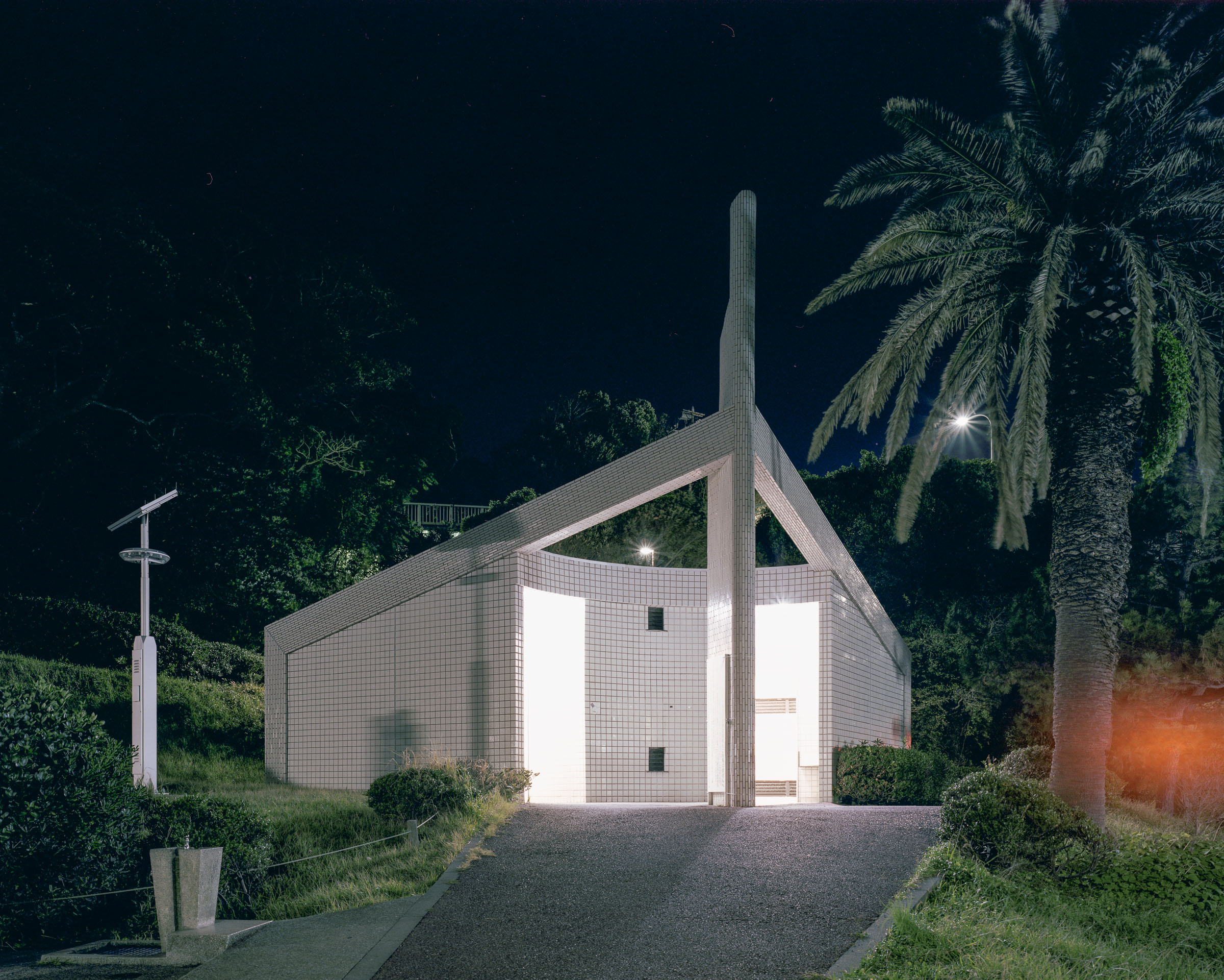 Angular tiled public toilet with a tall central column beside a palm tree