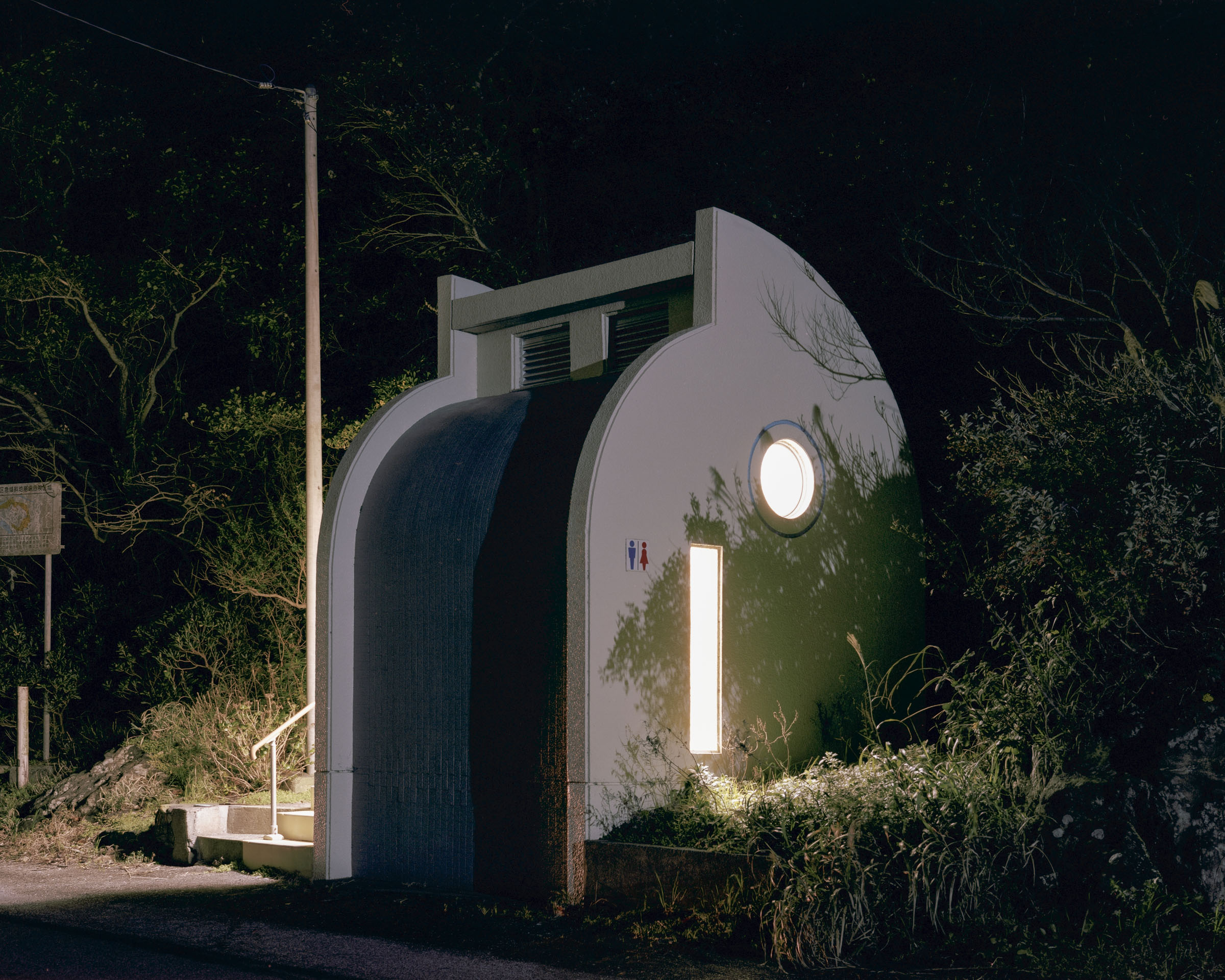 Curved white public toilet with round and vertical windows among trees