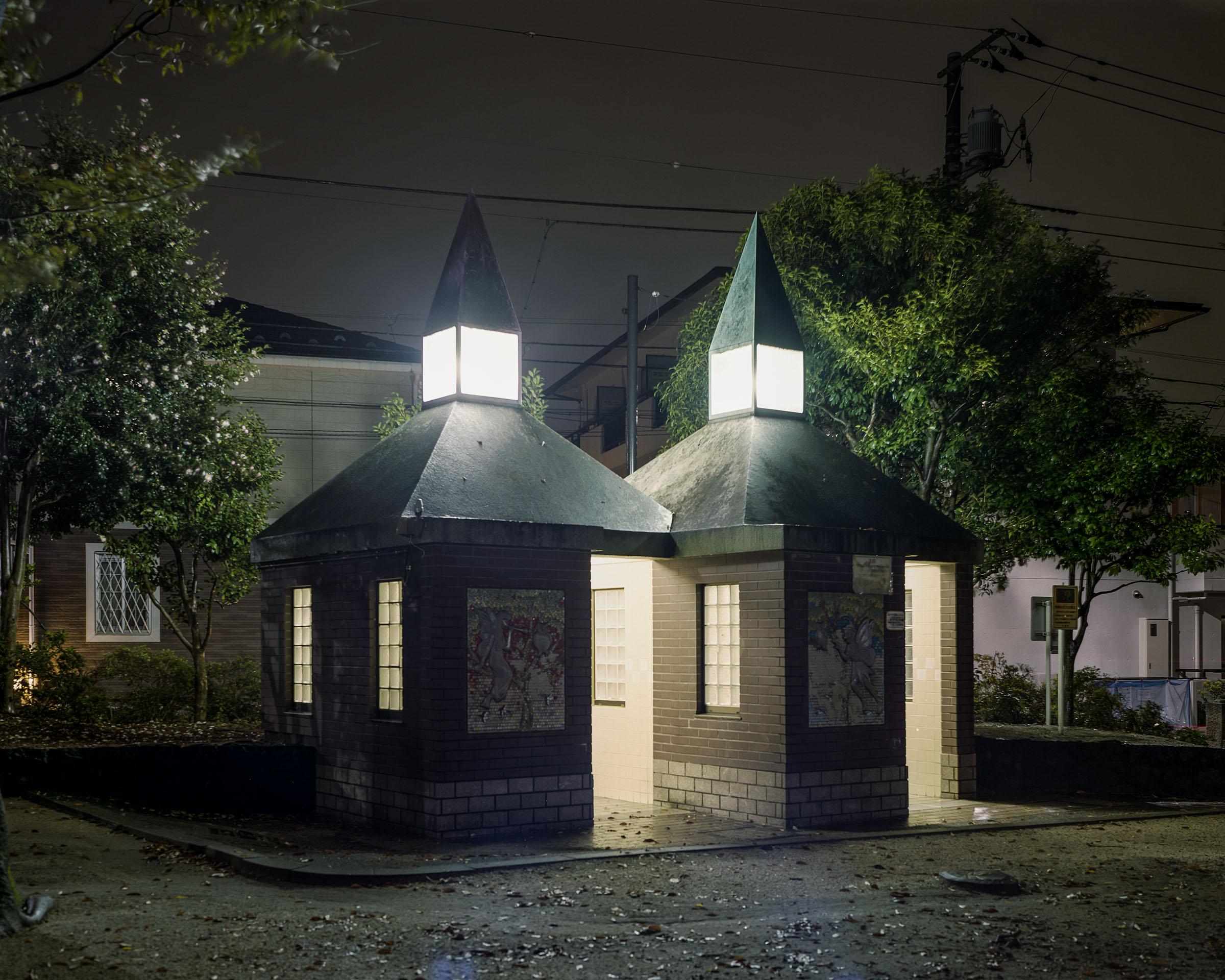 Twin brick toilet blocks with glowing pointed roof lanterns among trees